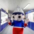 Mascot with a newspaper cone hat stands in a blue service hall, waving at the camera.