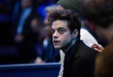 Close-up of a young man with dark hair in a black shirt, looking to the left in a dimly lit crowd.