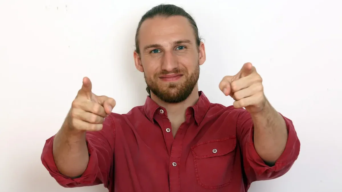 Smiling man in a red shirt points both index fingers at the camera.