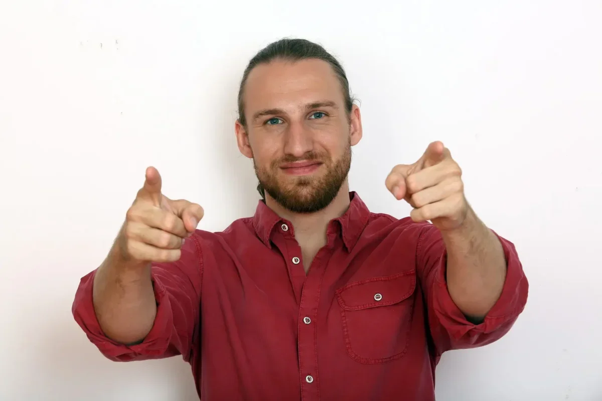 Smiling man in a red shirt points both index fingers at the camera.