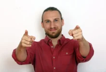 Smiling man in a red shirt points both index fingers at the camera.