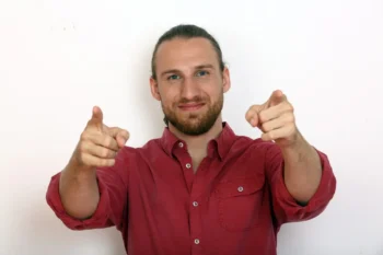 Smiling man in a red shirt points both index fingers at the camera.