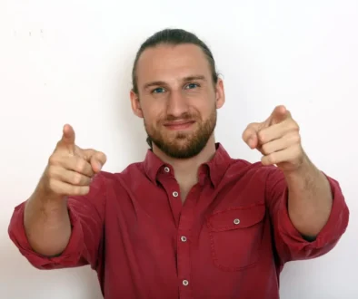 Smiling man in a red shirt points both index fingers at the camera.