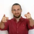 Smiling man in a red shirt points both index fingers at the camera.