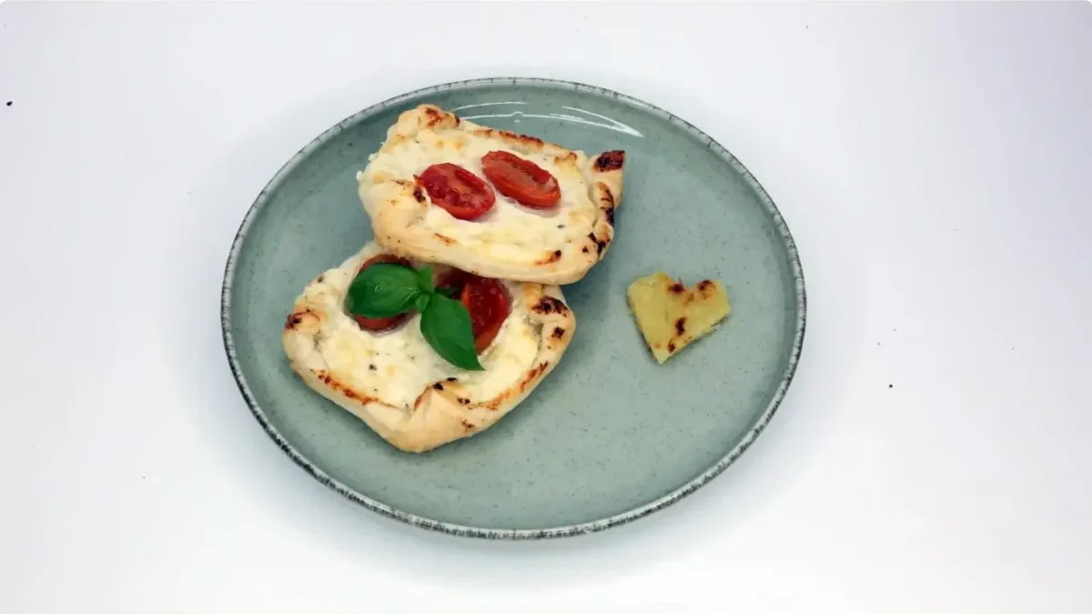 Top-down view of a gray plate with two mini flatbreads topped with cherry tomatoes and melted cheese, garnished with basil; a small heart-shaped bread piece rests on the side.