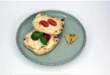 Top-down view of a gray plate with two mini flatbreads topped with cherry tomatoes and melted cheese, garnished with basil; a small heart-shaped bread piece rests on the side.