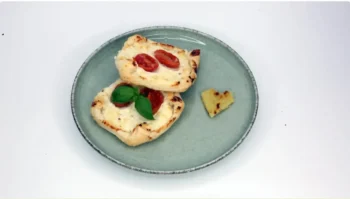 Top-down view of a gray plate with two mini flatbreads topped with cherry tomatoes and melted cheese, garnished with basil; a small heart-shaped bread piece rests on the side.