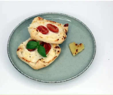 Top-down view of a gray plate with two mini flatbreads topped with cherry tomatoes and melted cheese, garnished with basil; a small heart-shaped bread piece rests on the side.