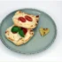 Top-down view of a gray plate with two mini flatbreads topped with cherry tomatoes and melted cheese, garnished with basil; a small heart-shaped bread piece rests on the side.