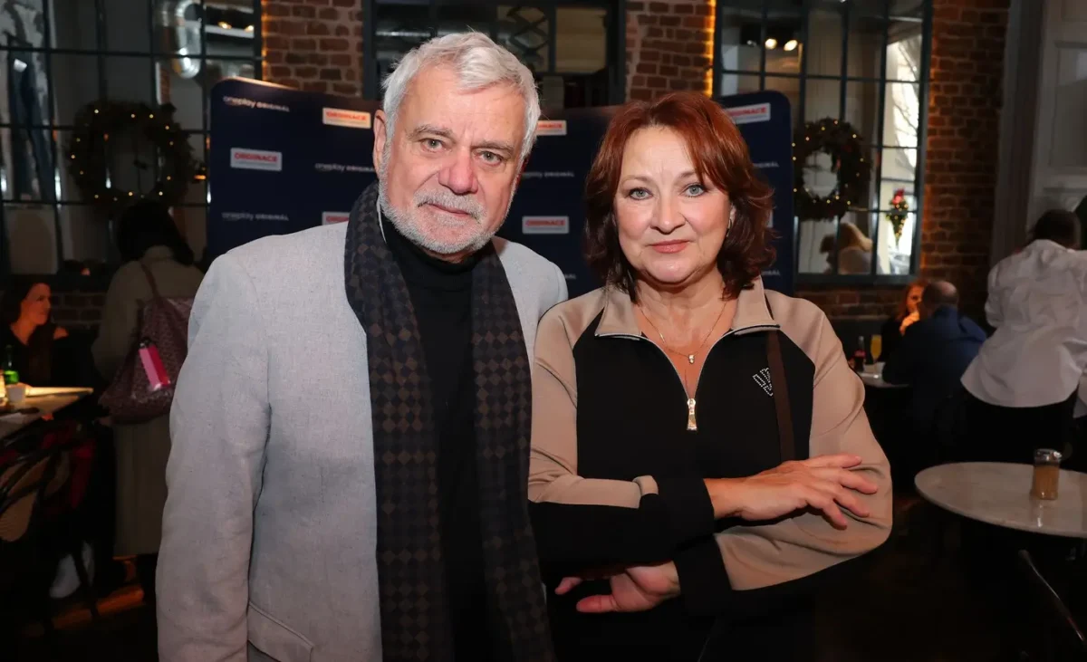 Two older adults posing together at a social event, smiling in front of a brick wall and backdrop decoration.