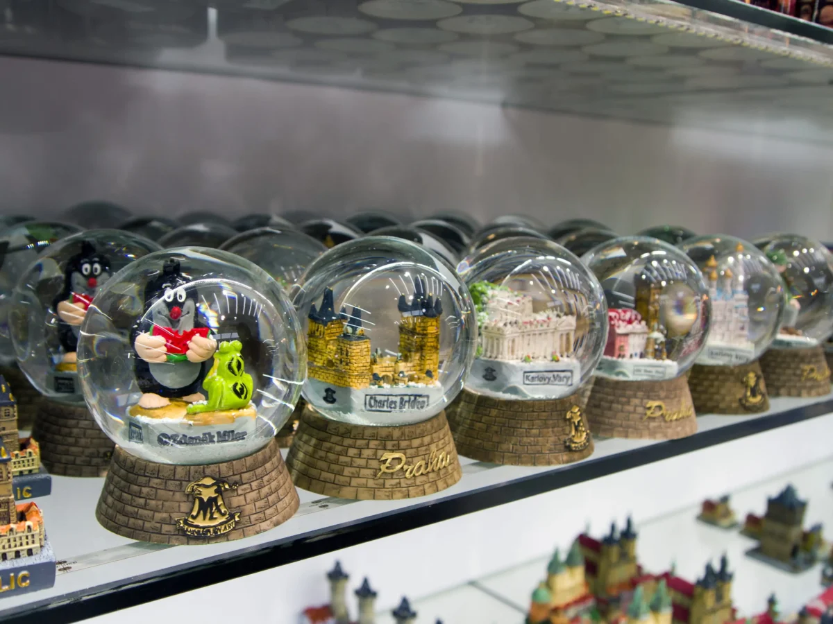 Row of glass snow globes on a store shelf, each containing miniature Prague landmarks and figures inside.