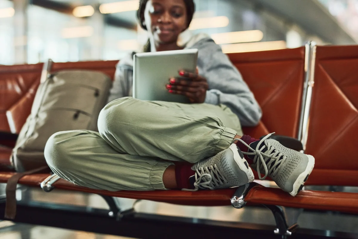 Person sitting on a red bench in a waiting area, cross-legged, holding a tablet, with a suitcase beside them in view.