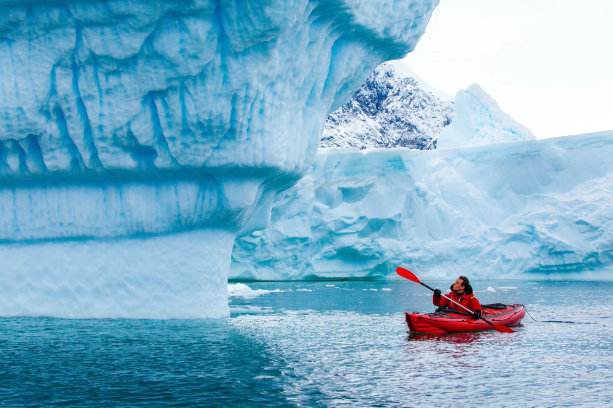 shutterstock_2720449499 1 | TVGURU.cz Kayaker in a red kayak paddling near towering blue iceberg formations in icy water