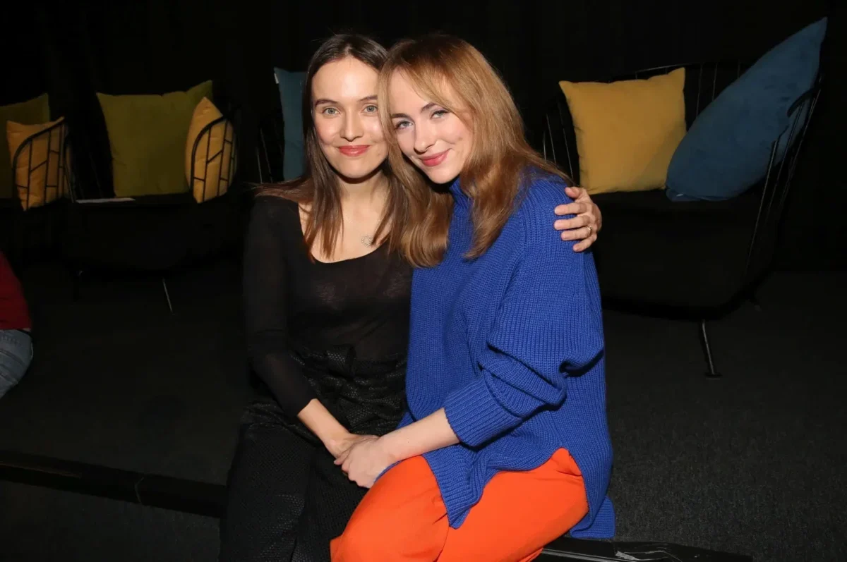Two women sit close together on a dark sofa, smiling at the camera; one wears a black top, the other a blue sweater and orange pants with colorful pillows behind them.