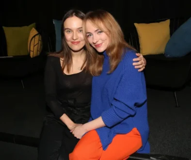 Two women sit close together on a dark sofa, smiling at the camera; one wears a black top, the other a blue sweater and orange pants with colorful pillows behind them.