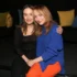 Two women sit close together on a dark sofa, smiling at the camera; one wears a black top, the other a blue sweater and orange pants with colorful pillows behind them.