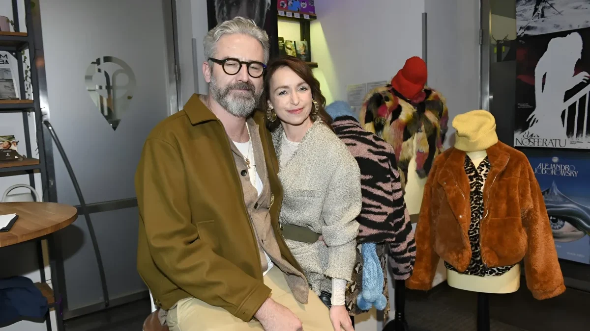 Man with gray hair and glasses and a smiling woman pose together in a fashion showroom, with colorful fur coats on mannequins in the background.