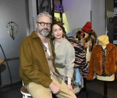 Man with gray hair and glasses and a smiling woman pose together in a fashion showroom, with colorful fur coats on mannequins in the background.