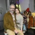 Man with gray hair and glasses and a smiling woman pose together in a fashion showroom, with colorful fur coats on mannequins in the background.