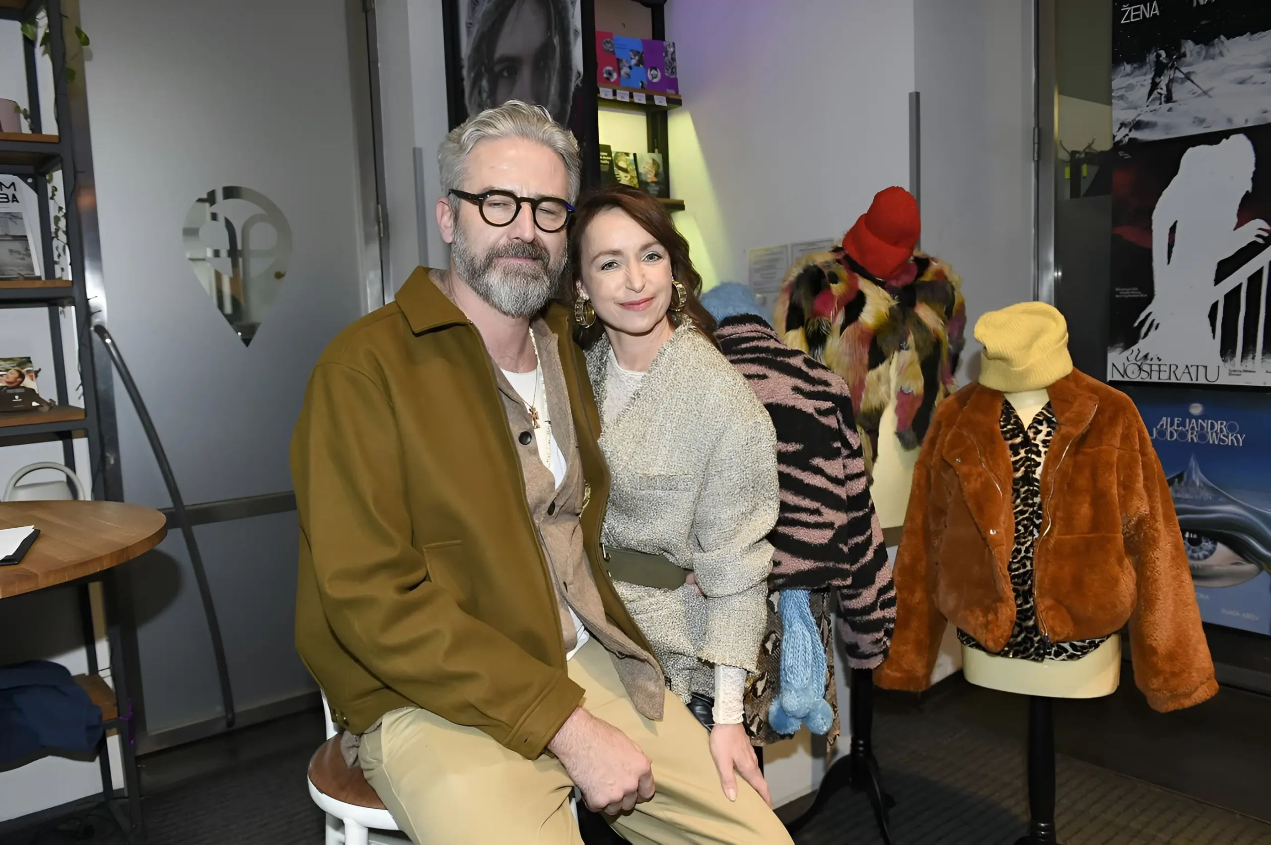 Man with gray hair and glasses and a smiling woman pose together in a fashion showroom, with colorful fur coats on mannequins in the background.
