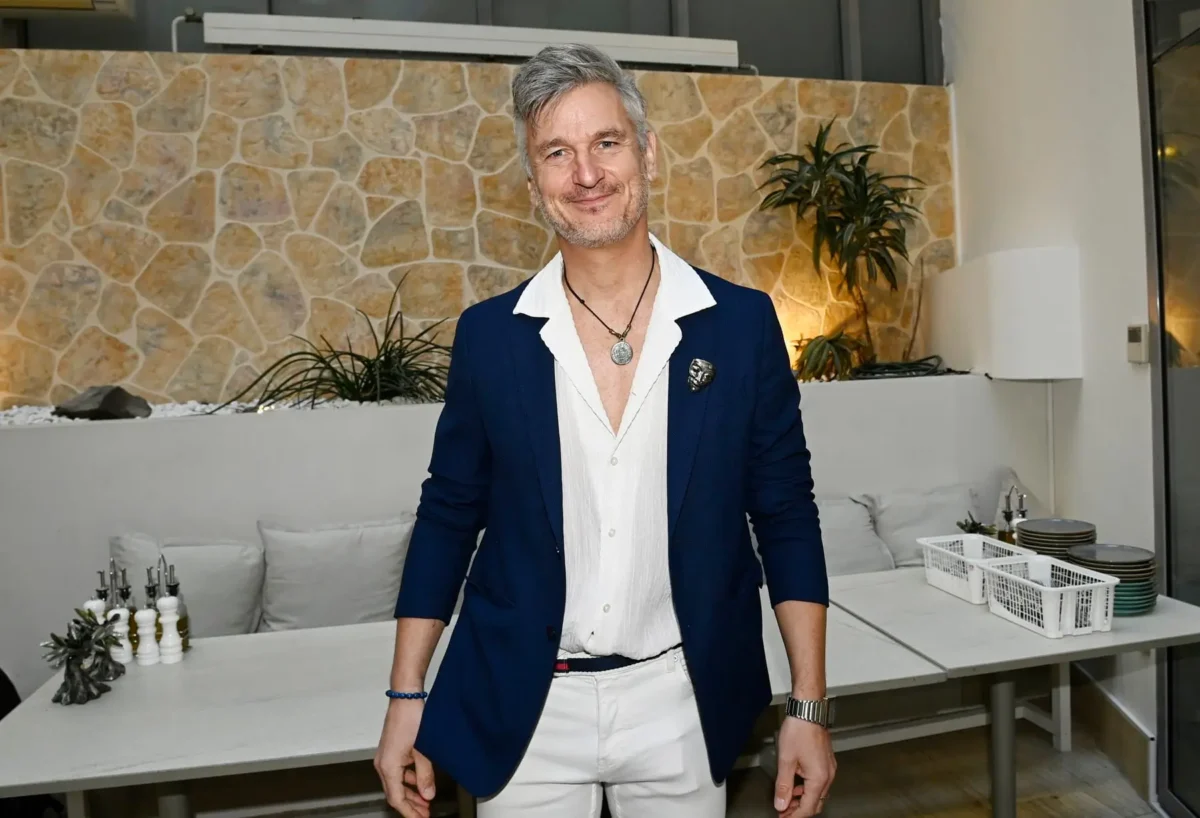 profimedia-1089366947 1 | TVGURU.cz Smiling man in a navy blazer and white shirt stands in a modern kitchen with a stone accent wall and plants behind him familiar dining area setting.