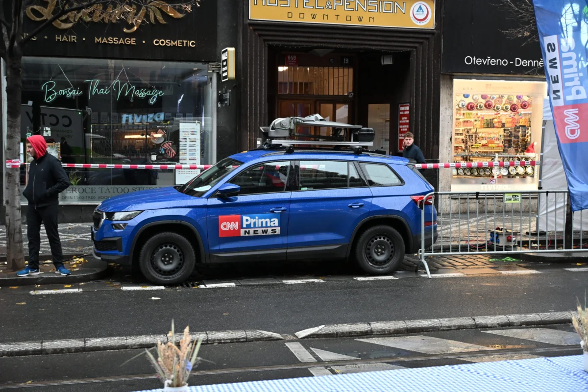 shutterstock_2706957117 1 | TVGURU.cz Blue CNN Prima News SUV parked on a city street with a roof rack, behind red-and-white barrier tape and storefronts.