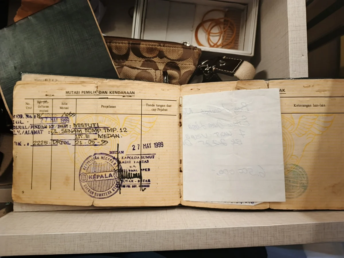 shutterstock_2809767135 1 | TVGURU.cz Open, aged Indonesian vehicle registration booklet with official stamp and handwritten notes in a wooden box shelf behind other items such as a brown patterned case and coiled cords.