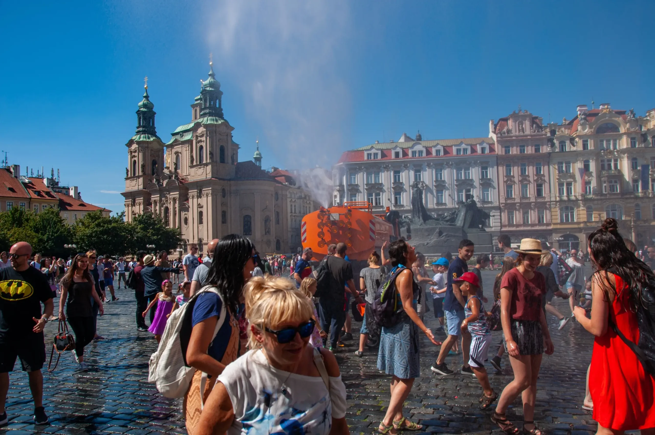 Lidé jsou v Praze na náměstí a kolem stříká voda.