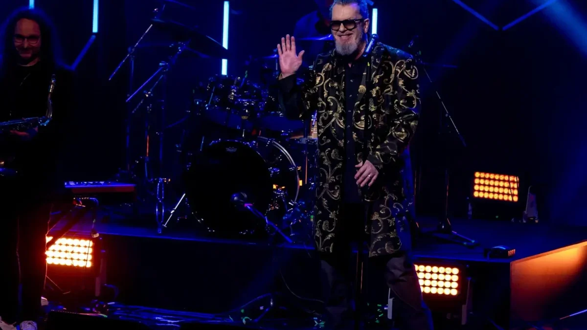 Smiling male singer in a gold-embroidered coat and sunglasses waves to the crowd on a dark stage, with a drummer behind him under blue lights.