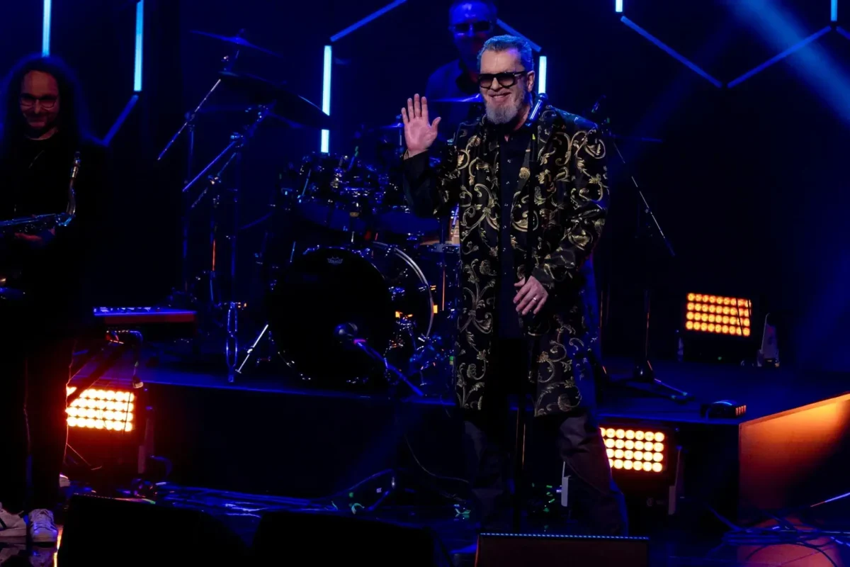 Smiling male singer in a gold-embroidered coat and sunglasses waves to the crowd on a dark stage, with a drummer behind him under blue lights.