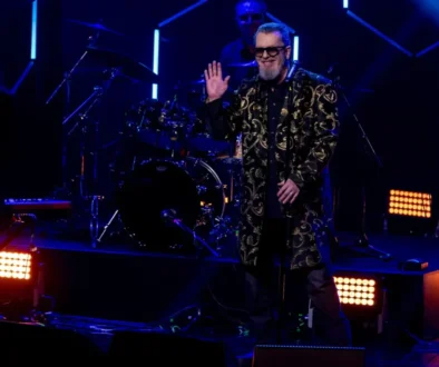 Smiling male singer in a gold-embroidered coat and sunglasses waves to the crowd on a dark stage, with a drummer behind him under blue lights.
