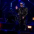 Smiling male singer in a gold-embroidered coat and sunglasses waves to the crowd on a dark stage, with a drummer behind him under blue lights.
