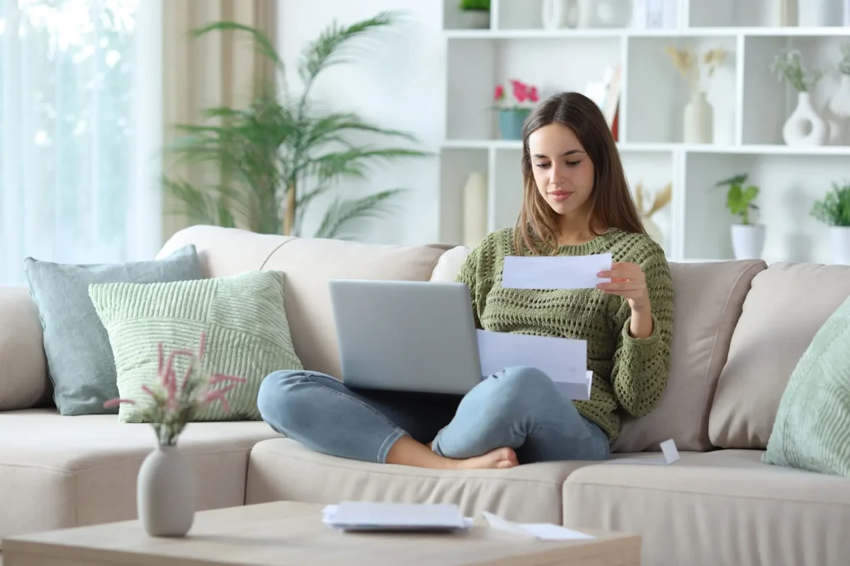 shutterstock_2724840049 1 | TVGURU.cz mladá žena sedí na gauči s notebookem na klíně a pročítá papírový dopis