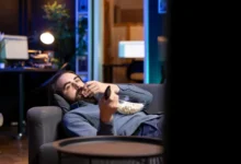 muž leží na gauči s ovladačem a popcornem a kouká na televizi