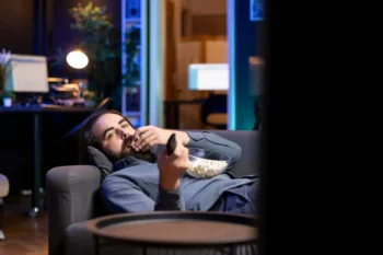 muž leží na gauči s ovladačem a popcornem a kouká na televizi
