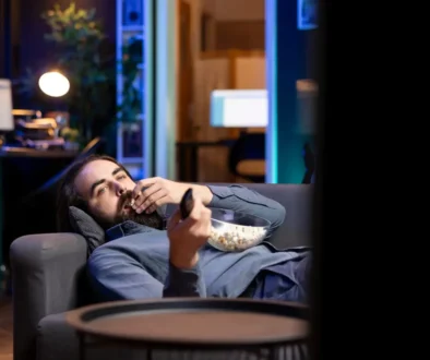 muž leží na gauči s ovladačem a popcornem a kouká na televizi