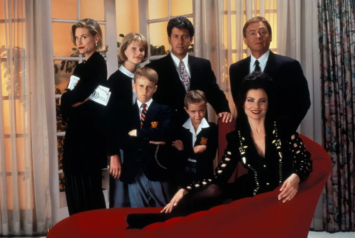 A group of adults and children in formal attire posing in a bright room, with a woman in a shiny black jacket seated on a red sofa in front.