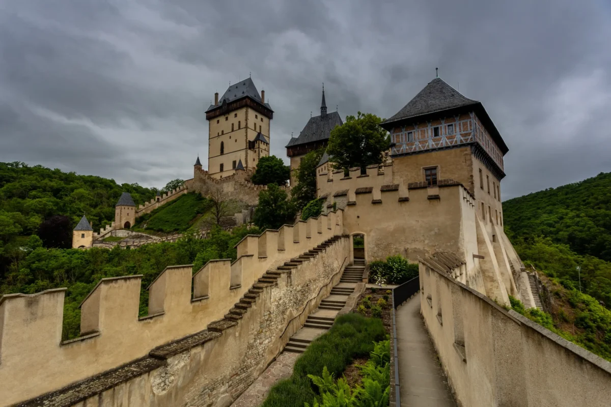 Středověká pevnost Karlštejn na kopci s béžovými kamennými zdmi, věžemi a točitými schodišti pod zataženou oblohou.