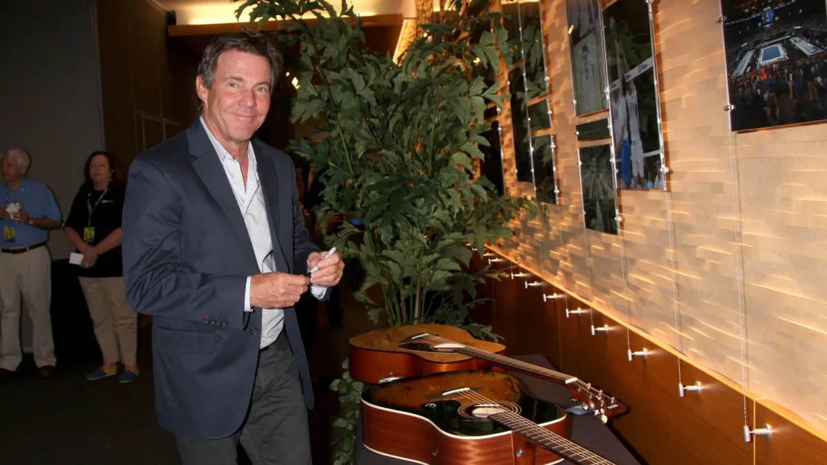 Man in a navy blazer signs a guitar at a display wall in a showroom, with potted plants behind him and photos on the wall to the right.