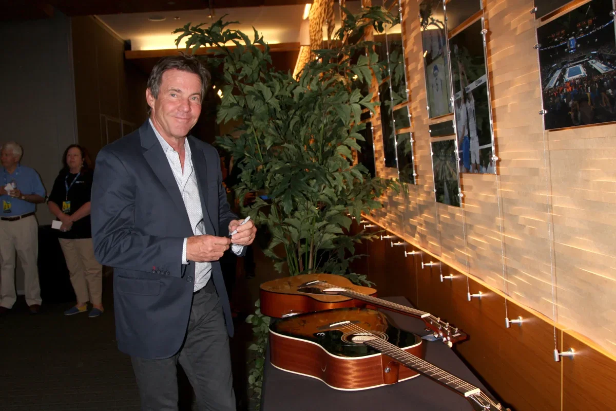 Man in a navy blazer signs a guitar at a display wall in a showroom, with potted plants behind him and photos on the wall to the right.