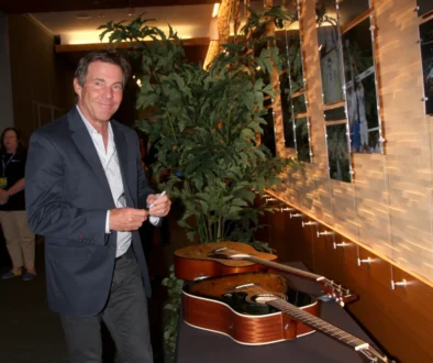 Man in a navy blazer signs a guitar at a display wall in a showroom, with potted plants behind him and photos on the wall to the right.