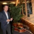Man in a navy blazer signs a guitar at a display wall in a showroom, with potted plants behind him and photos on the wall to the right.