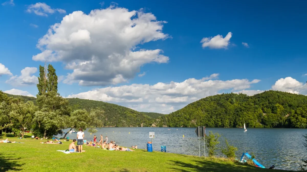 People sunbathing on a grassy lakeshore with a blue sky and clouds, and tree-covered hills in the background.