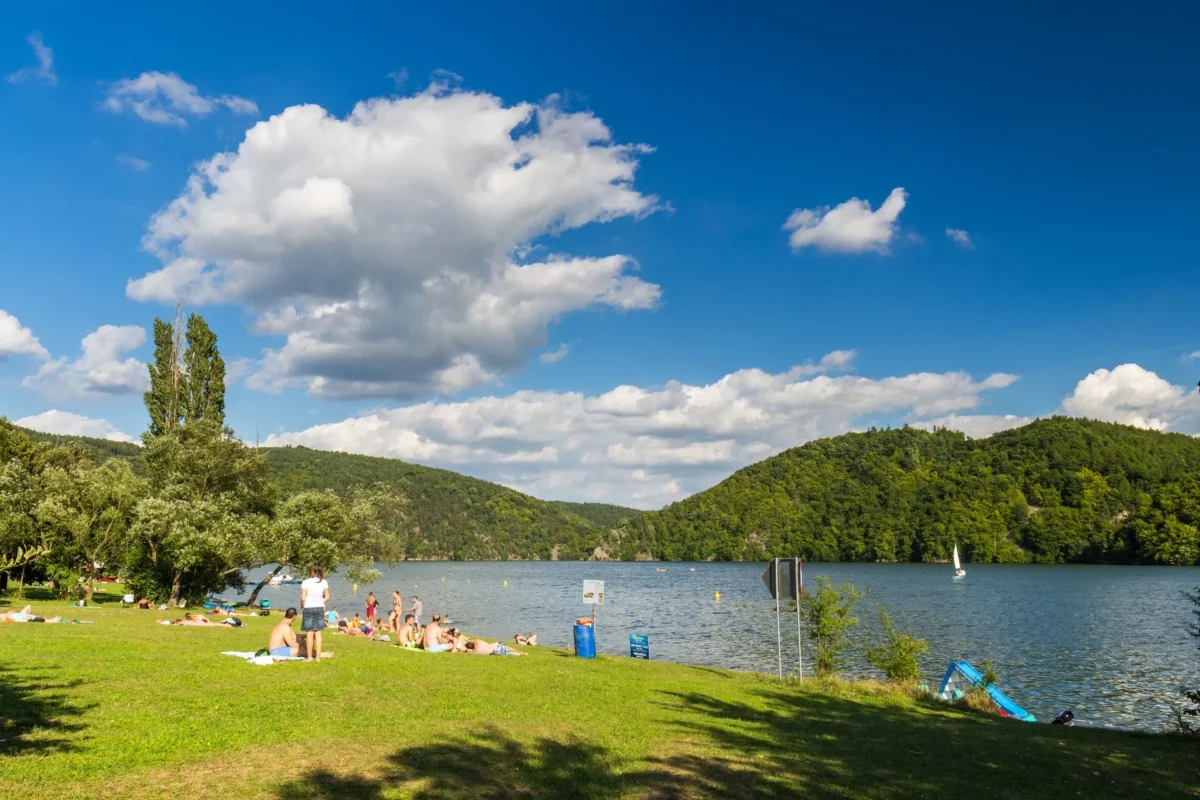People sunbathing on a grassy lakeshore with a blue sky and clouds, and tree-covered hills in the background.