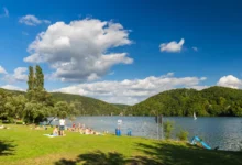 People sunbathing on a grassy lakeshore with a blue sky and clouds, and tree-covered hills in the background.