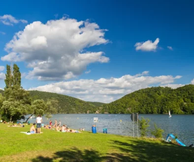 People sunbathing on a grassy lakeshore with a blue sky and clouds, and tree-covered hills in the background.