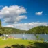 People sunbathing on a grassy lakeshore with a blue sky and clouds, and tree-covered hills in the background.