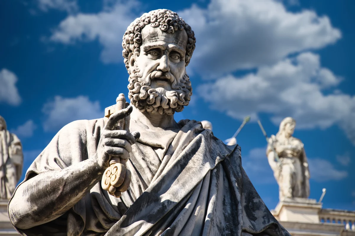 shutterstock_2442281553 1 | TVGURU.cz Stone statue of a bearded man in robes, holding a staff, against a blue sky with clouds.