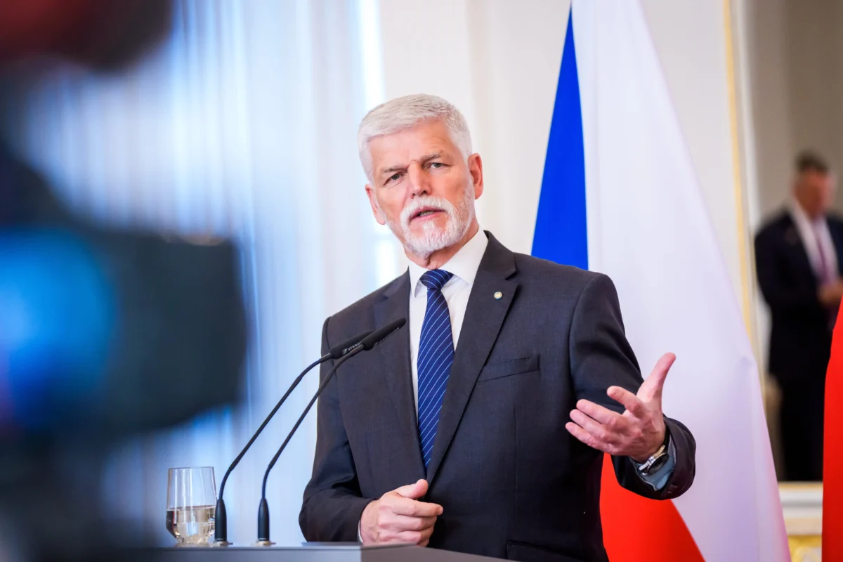 shutterstock_2752187265 1 | TVGURU.cz Man in a dark suit and blue striped tie speaks at a podium with microphones, flags in the background.