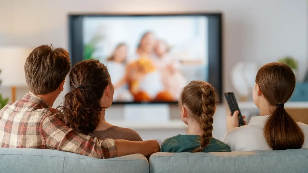 Family seated on a sofa watching a blurry TV show together, one person holding a remote control to the right